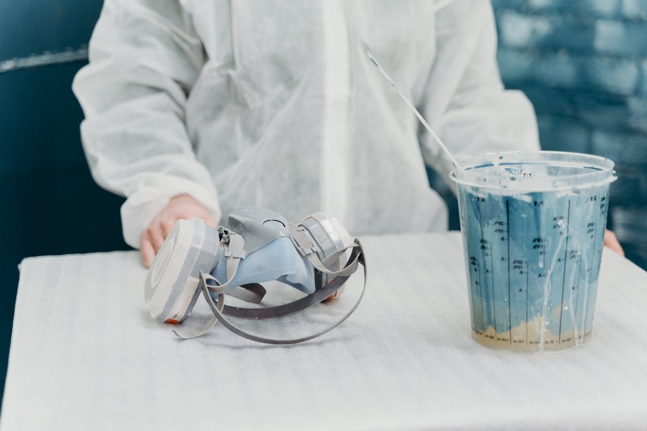 A person in protective gear standing near a gas mask and paint can indoors.